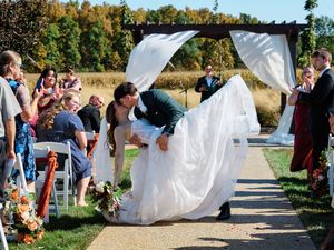 White wedding ceremony setup outdoors with flowing fabric and chairs along a path surrounded by autumn trees.