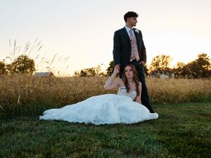Wedding party sits together on grass at sunset with a flowing white dress in view.