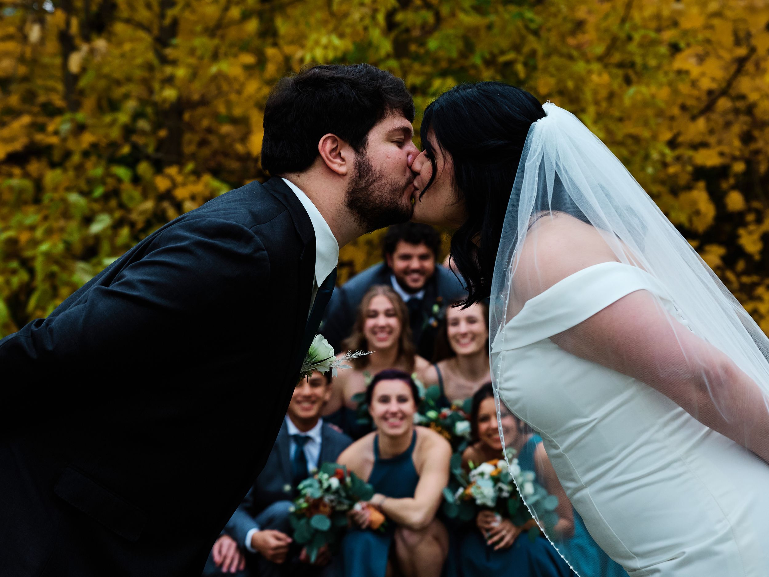 A romantic outdoor wedding scene with a couple embracing while guests smile and celebrate in the autumn foliage.