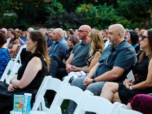 A crowd of people sits in white folding chairs at an outdoor event during summer.