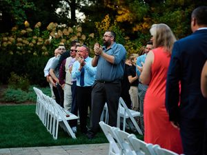 Wedding guests gather around white chairs set up outdoors for a garden ceremony at sunset.