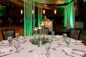 Elegant wedding reception tables set with white linens, green uplighting, and tall glass centerpiece vases.