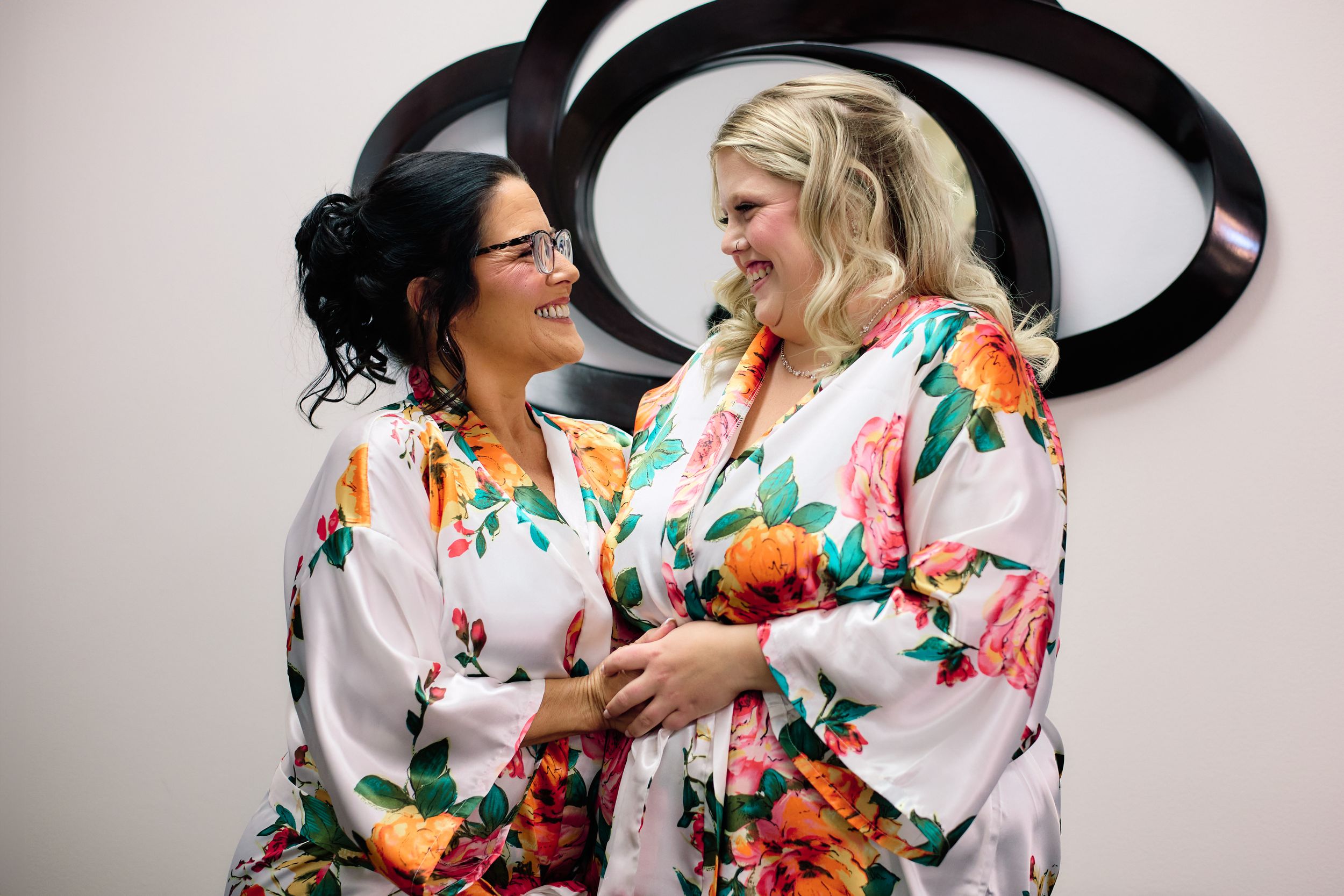 Two friends share a joyful moment wearing matching floral robes against a wall with a black logo.