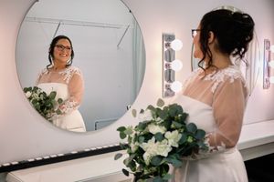A bride in a white dress holds a green and white bouquet while looking at her reflection in a dressing room mirror.