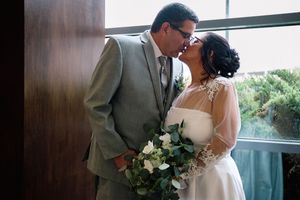 A couple shares an intimate moment by a window while holding a white and green floral bouquet.