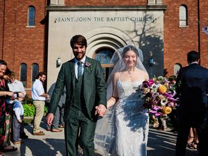 Wedding party members in green suits and white dresses exit a brick church building as guests celebrate with confetti.