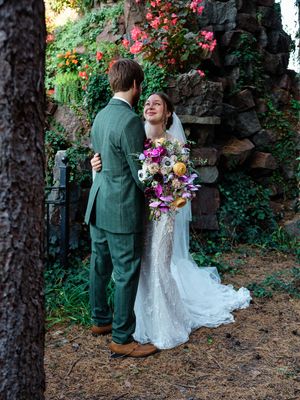 A joyful wedding couple shares a moment in a garden with blooming flowers and stone wall backdrop.