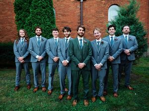 A group of groomsmen in matching gray suits pose together against a brick wall with green hedges.