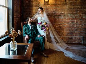 A wedding couple poses in a rustic brick-walled room with natural window light and flowing veil.