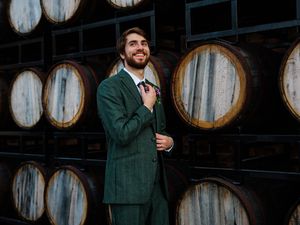 Wedding party members in dark green suits posing near wooden wine barrels in a rustic venue.