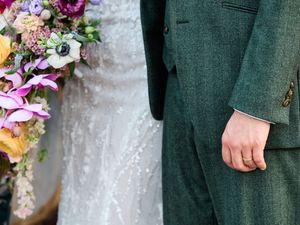 Close-up of vibrant purple and pink wedding bouquet against dark green formal attire.