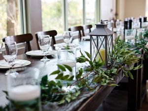 Long wedding reception table decorated with white linens, glassware and lush greenery garlands in an elegant venue.