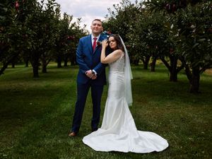 A couple poses for a wedding photo in a lush apple orchard wearing a blue suit and flowing white gown.
