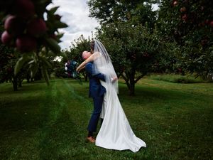 A couple in wedding attire dances together in a lush green apple orchard on their special day.