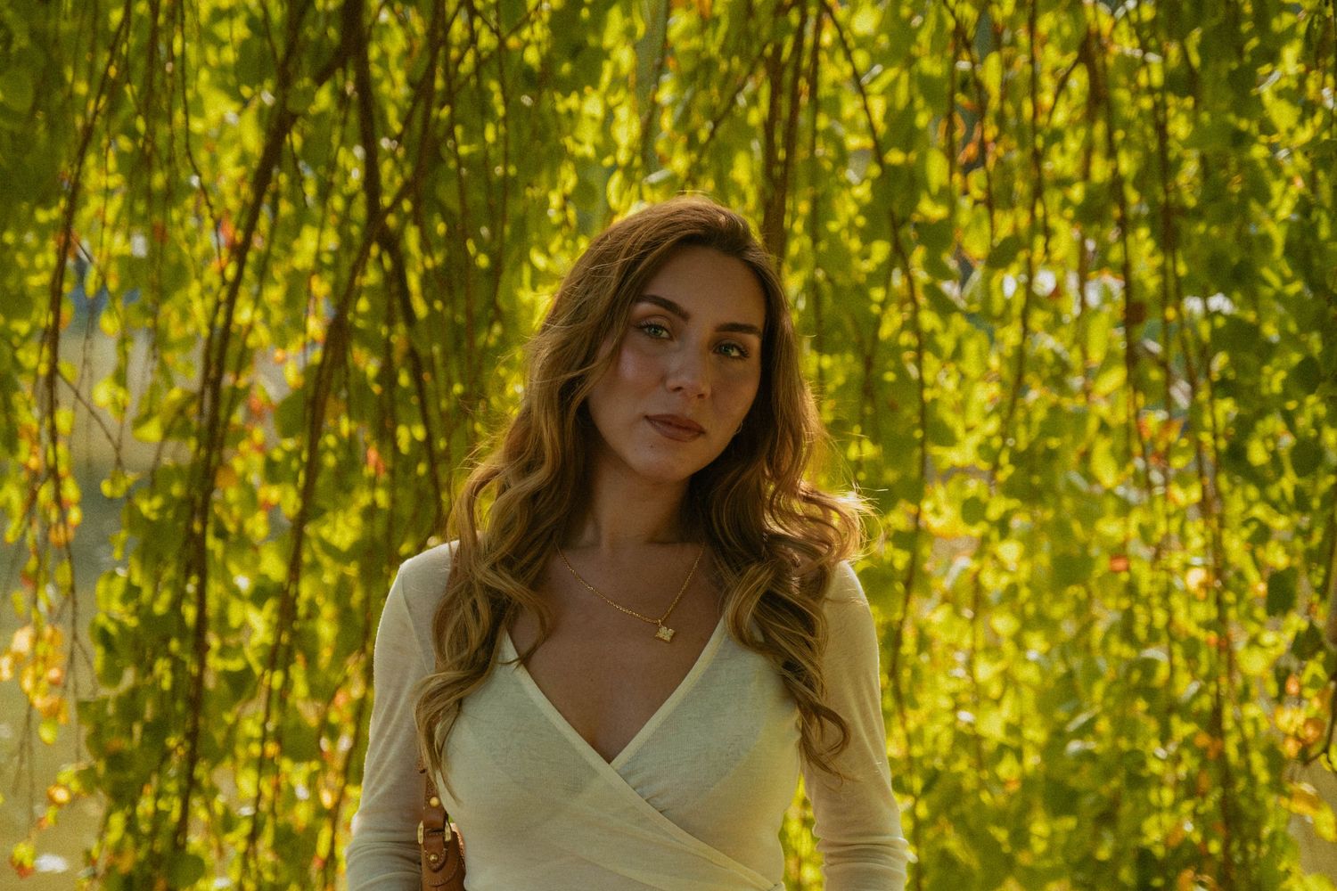 A woman poses under golden willow branches in natural sunlight.