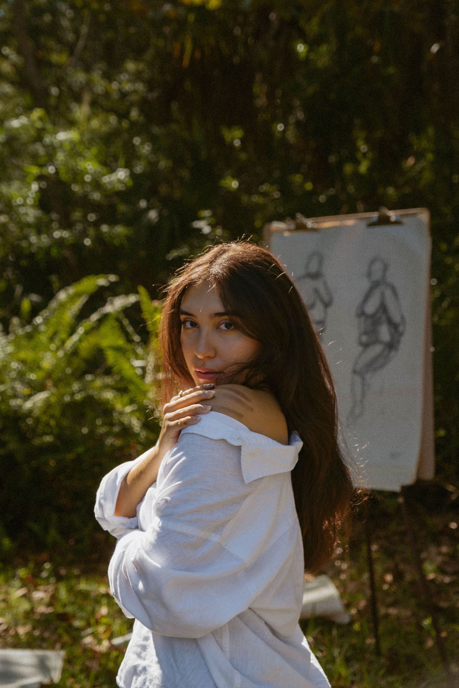Person in white button-down shirt poses artistically against natural outdoor backdrop with sketches visible in background.