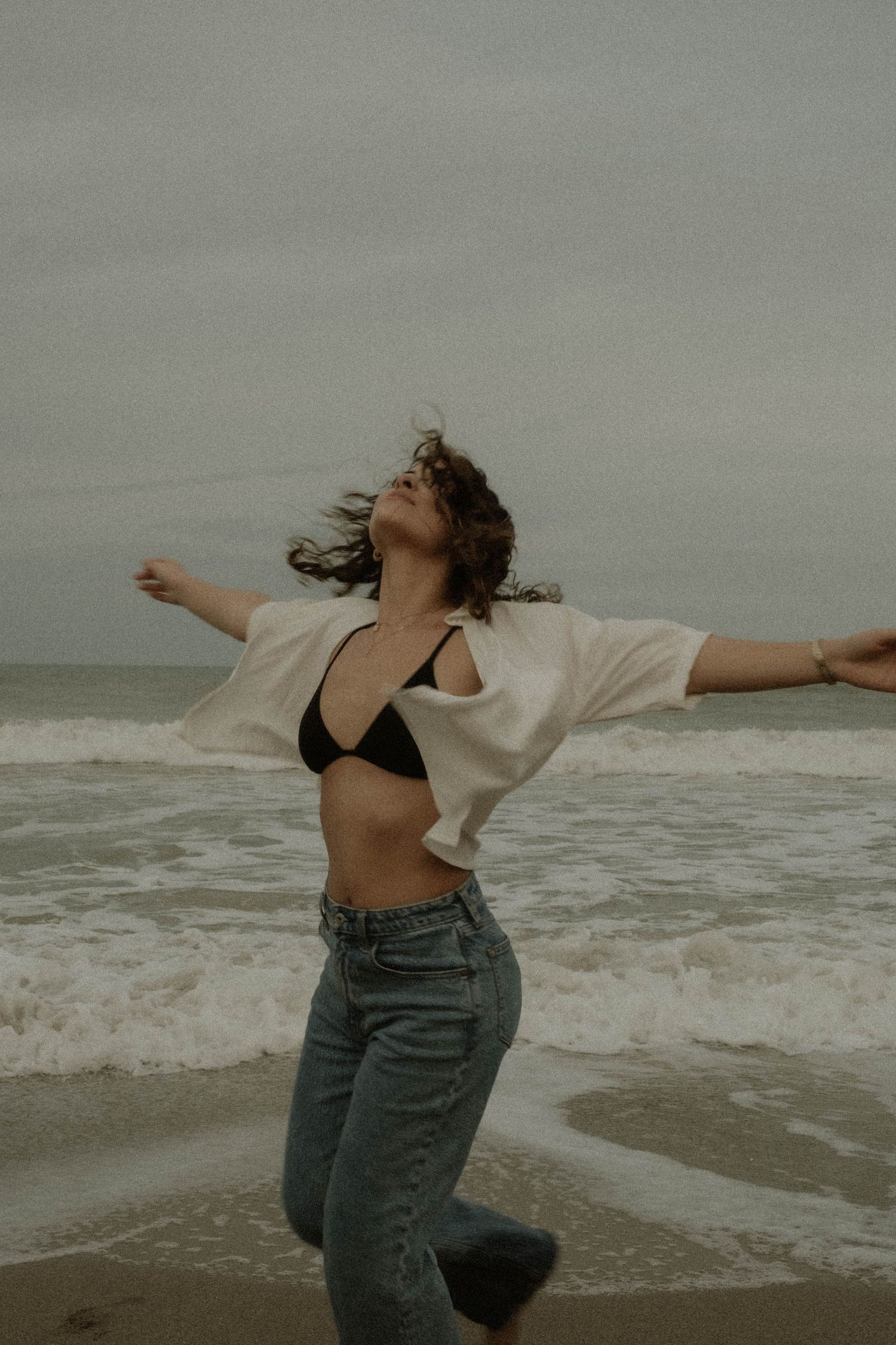 Figure with curly hair dances freely on a moody beach wearing jeans and an open shirt.