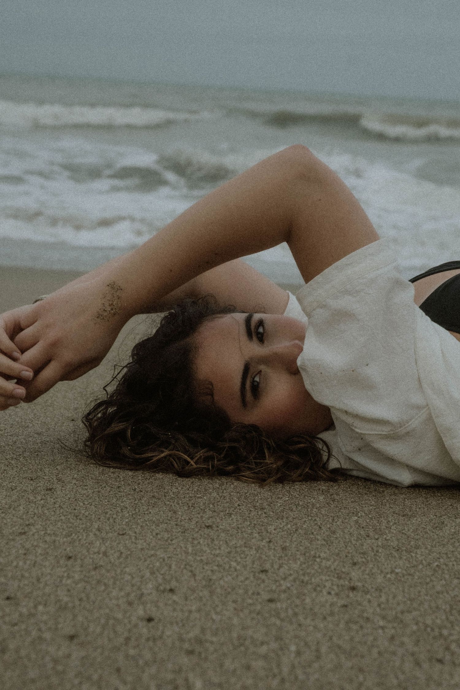 Person lying on sandy beach with waves in background during overcast day.