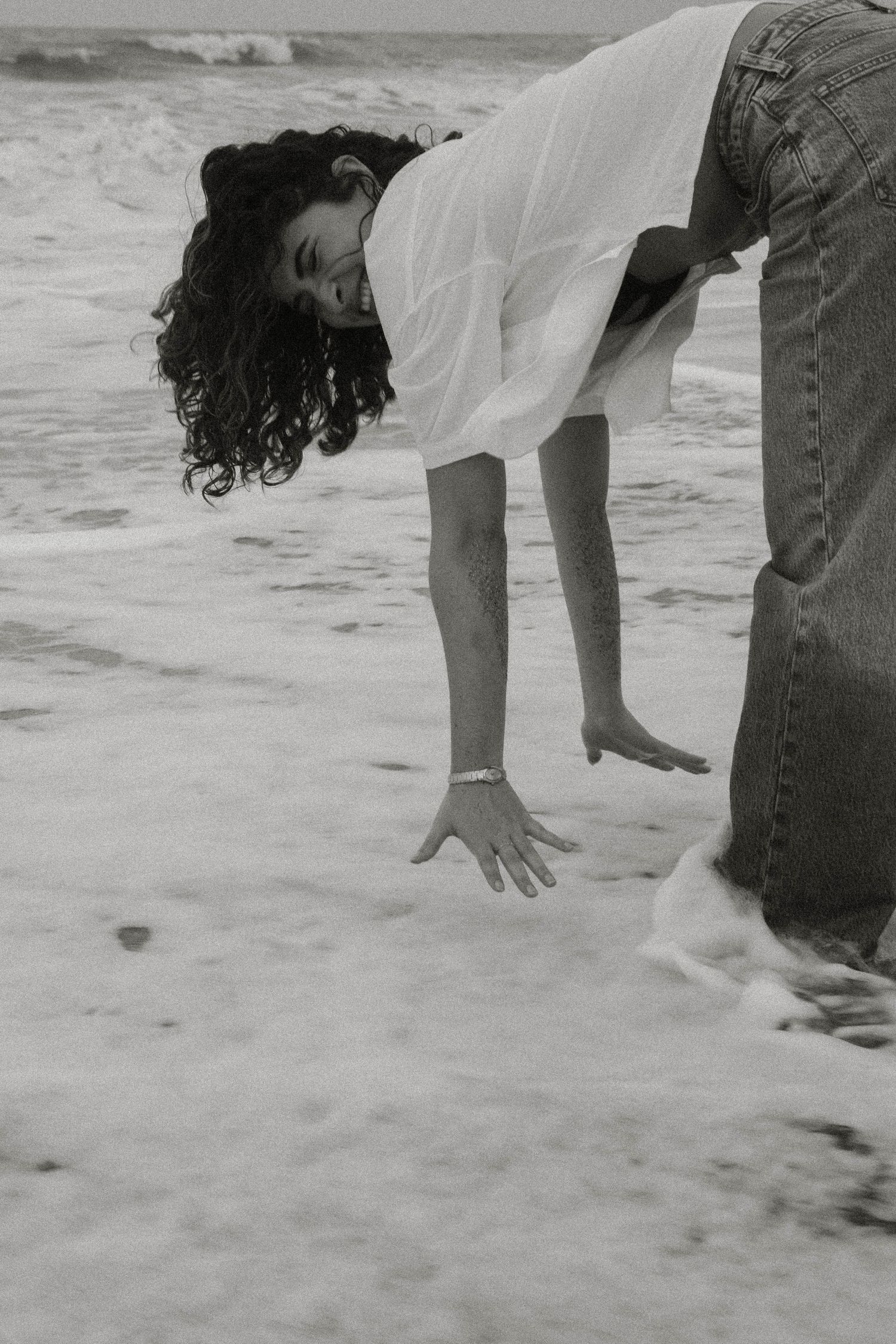 Person playing in ocean waves bends down to touch the water on a cloudy beach.