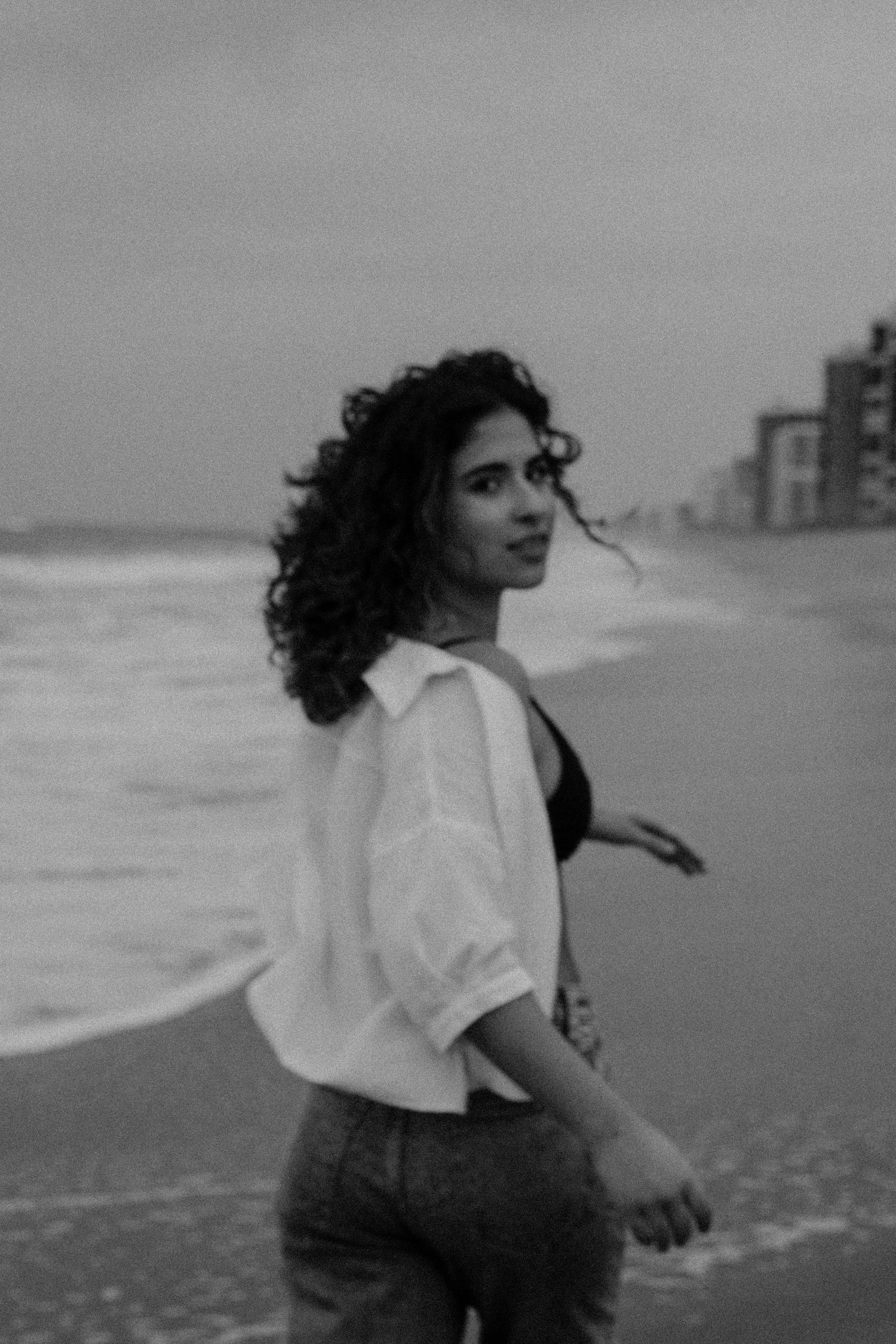 Artistic black and white photo of someone dancing carefree on a beach with buildings in distance.