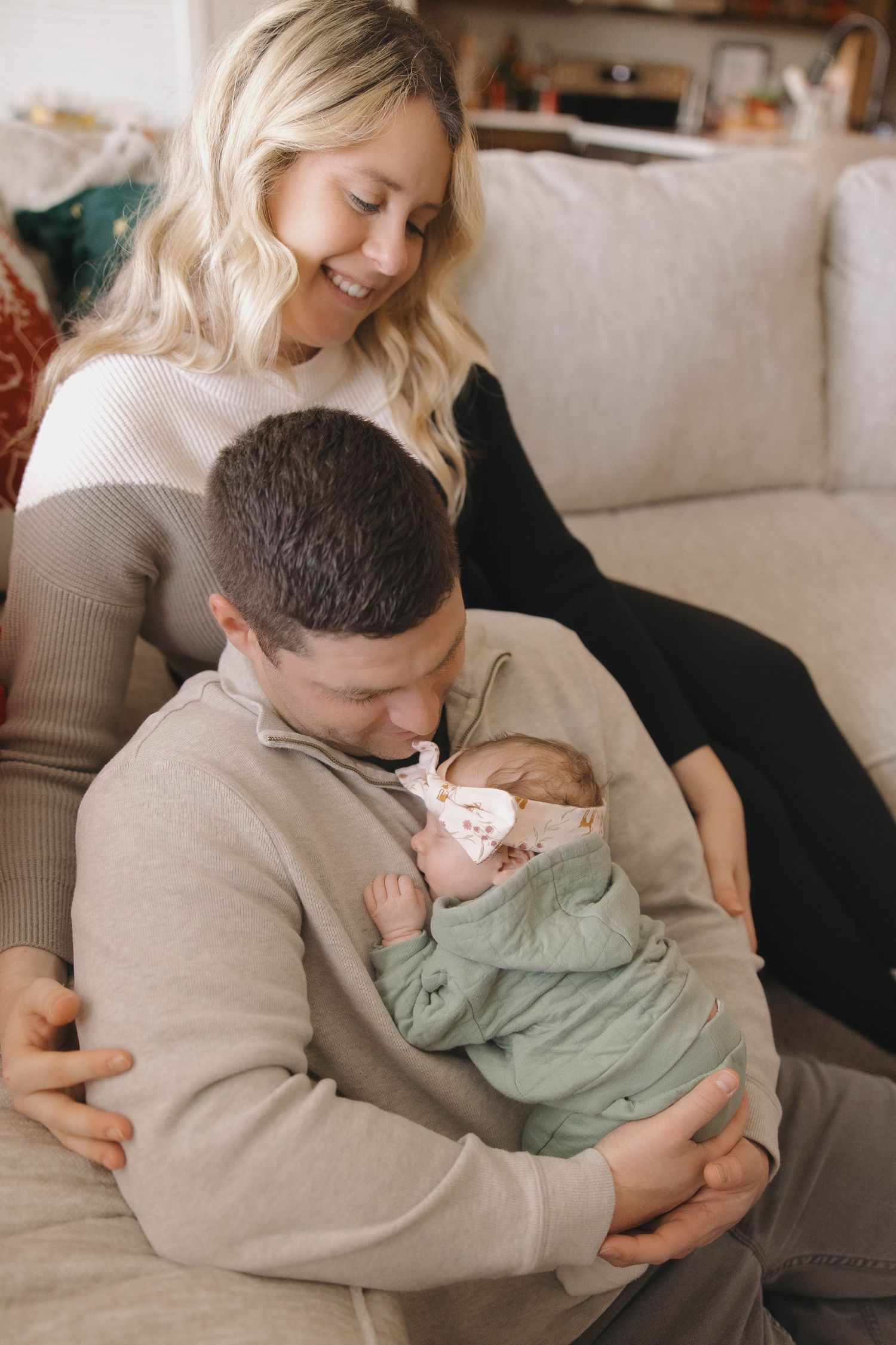 A tender family moment as parents cuddle with their newborn baby wearing a mint green outfit on a cozy couch.