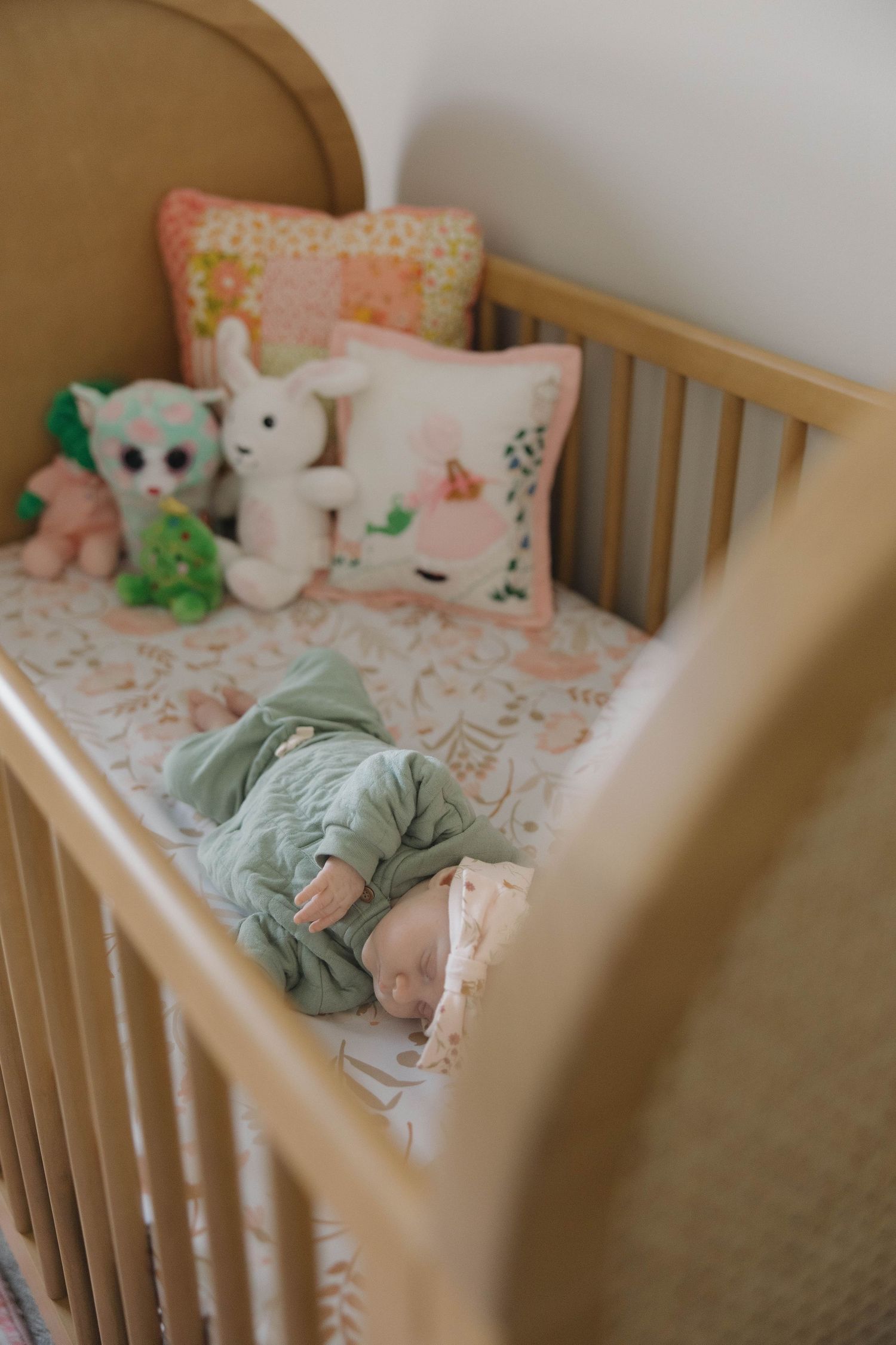 A wooden crib with pink floral bedding and plush toys arranged neatly against a light colored wall.