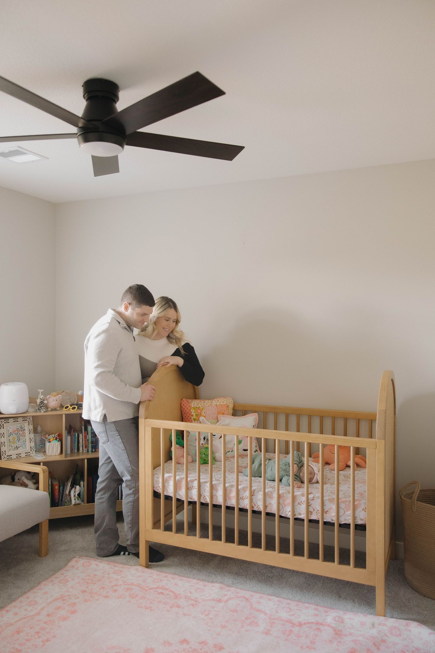 A wooden crib sits in a neutral-toned nursery with a ceiling fan and natural lighting.