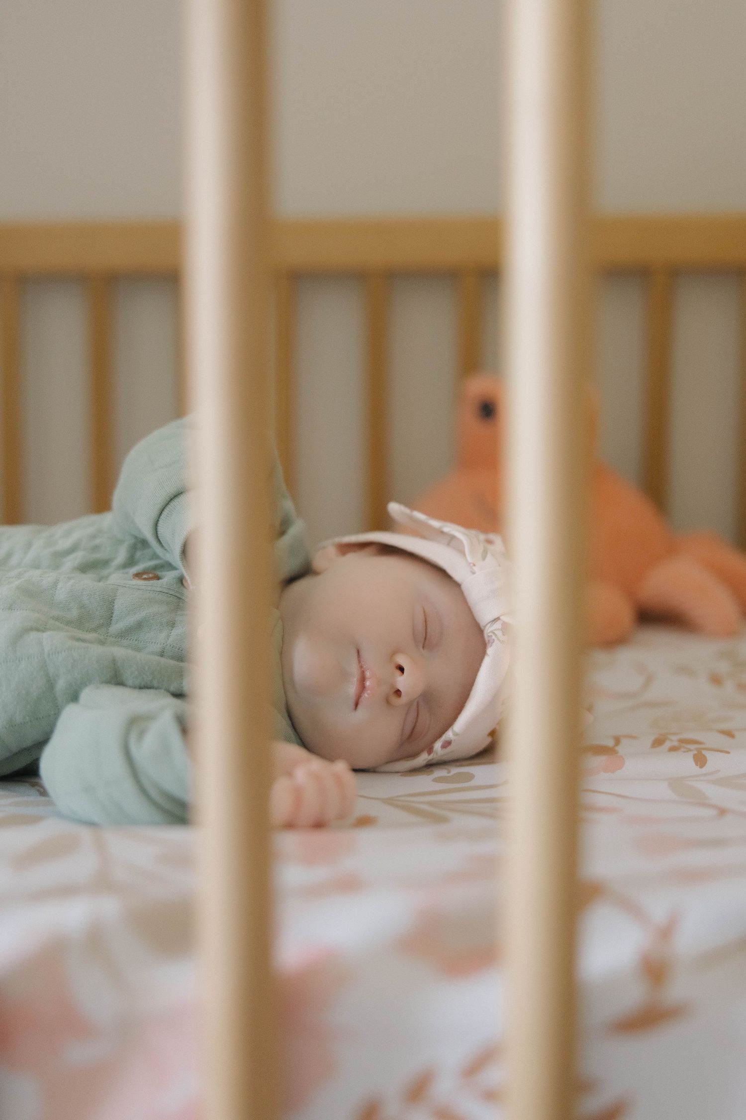 A sleeping baby in a white crib wearing a mint green outfit rests peacefully on floral bedding.