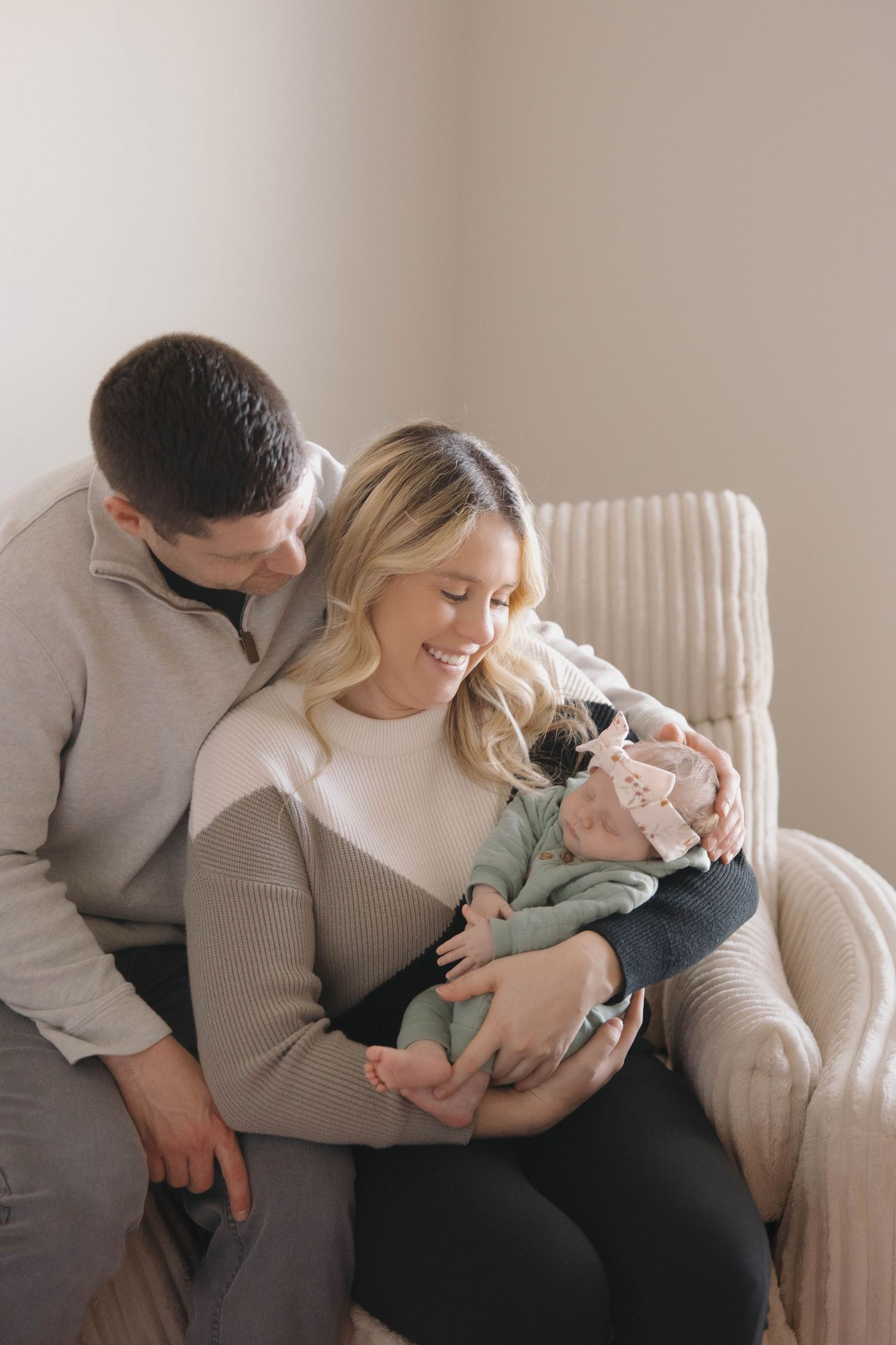 A couple snuggles with their baby on a white armchair in a tender family moment.