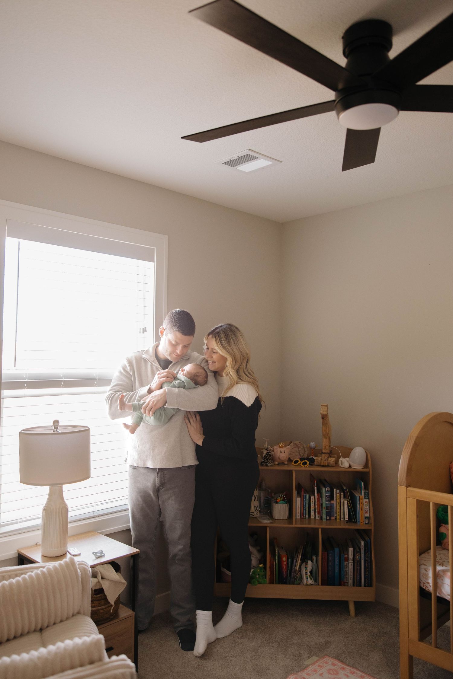 Parents sharing a special moment with their newborn in a nursery with bookshelves and ceiling fan.
