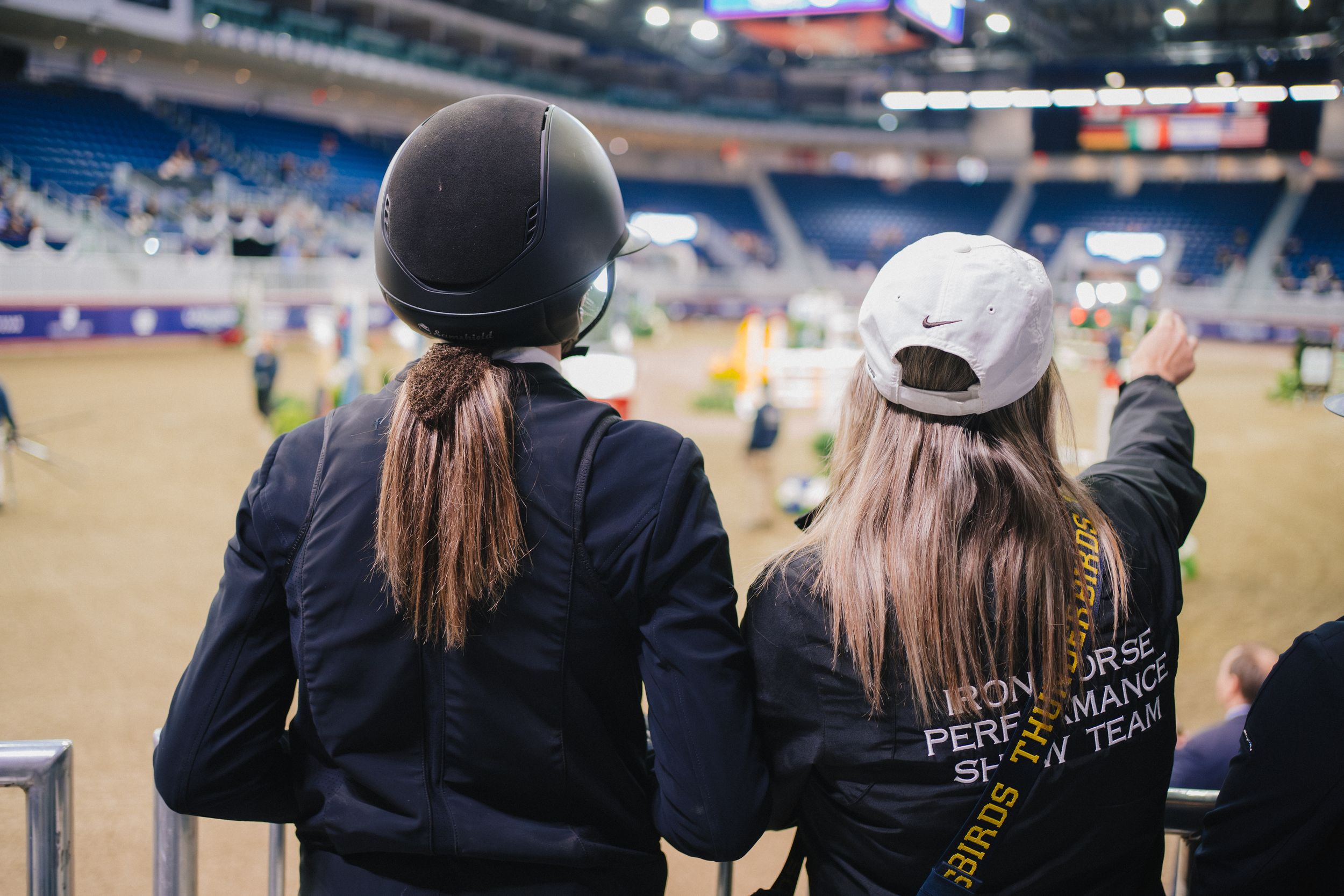 Equestrian riders in dark jackets and helmets watching from the sidelines at an indoor arena competition.