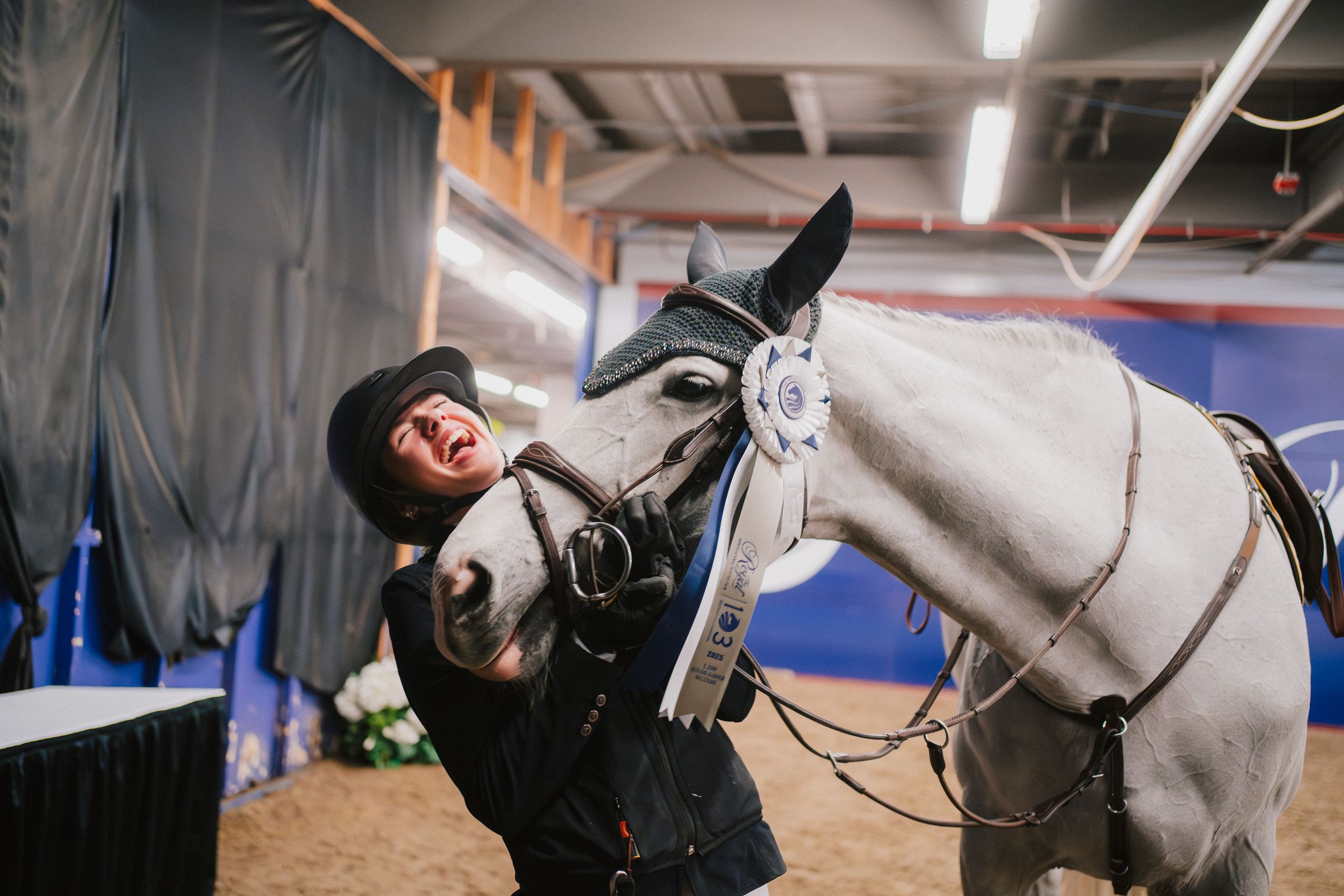 A white horse and rider practicing dressage movements in an indoor arena with bright lighting.