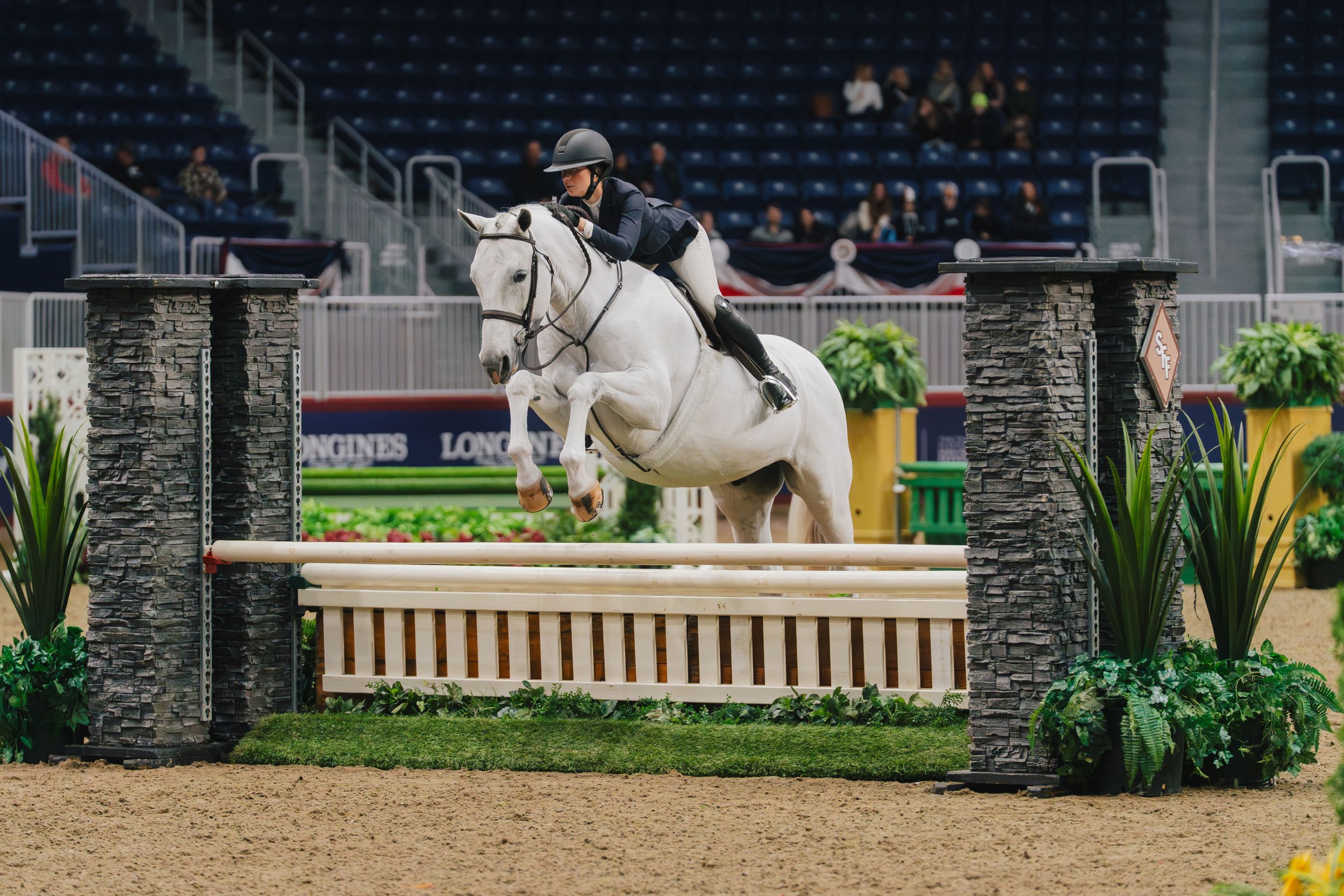 A horse and rider jump over a stone wall obstacle during an equestrian show jumping competition.