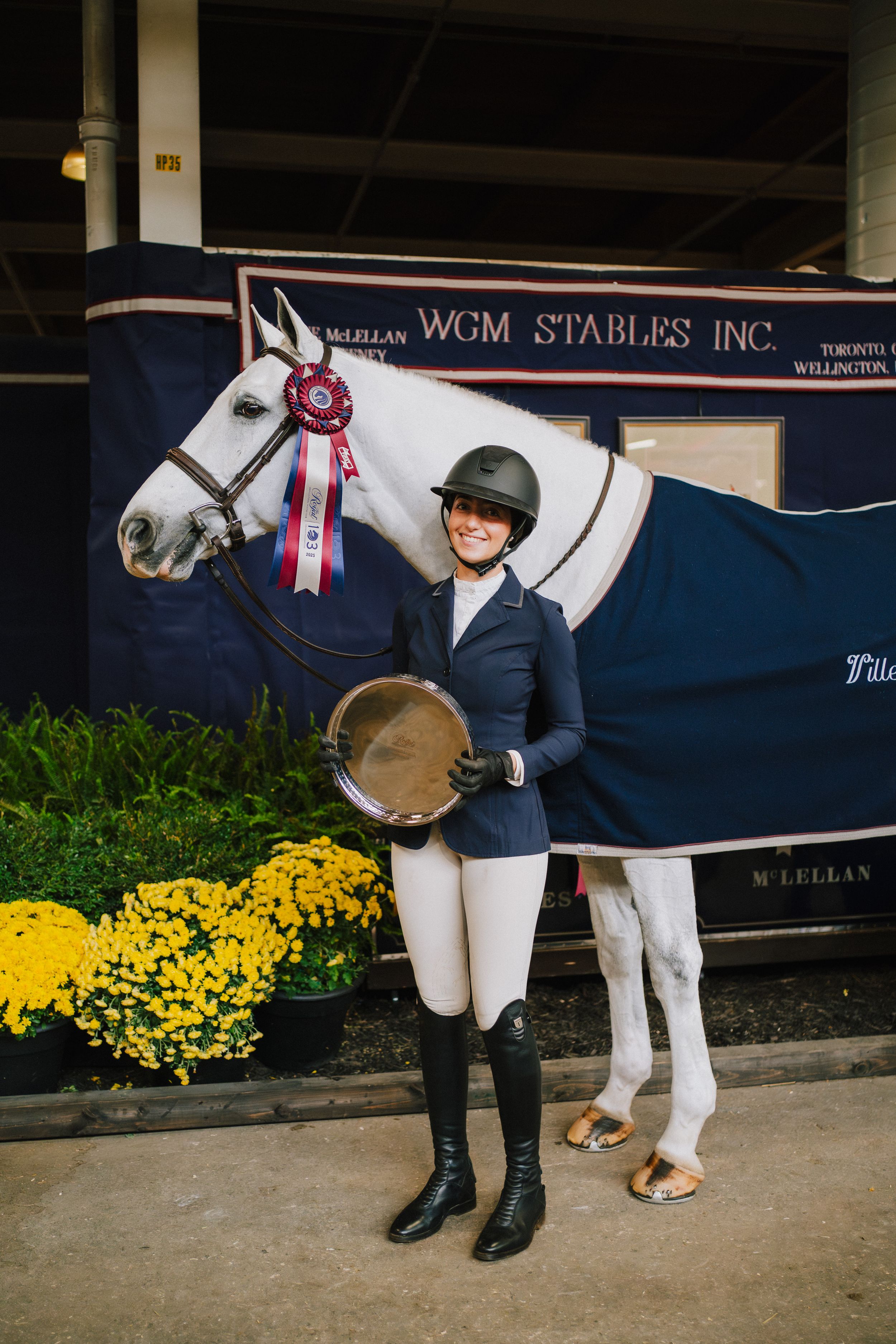 Equestrian competition participants stand with their horses in front of yellow flowers and display ribbons.