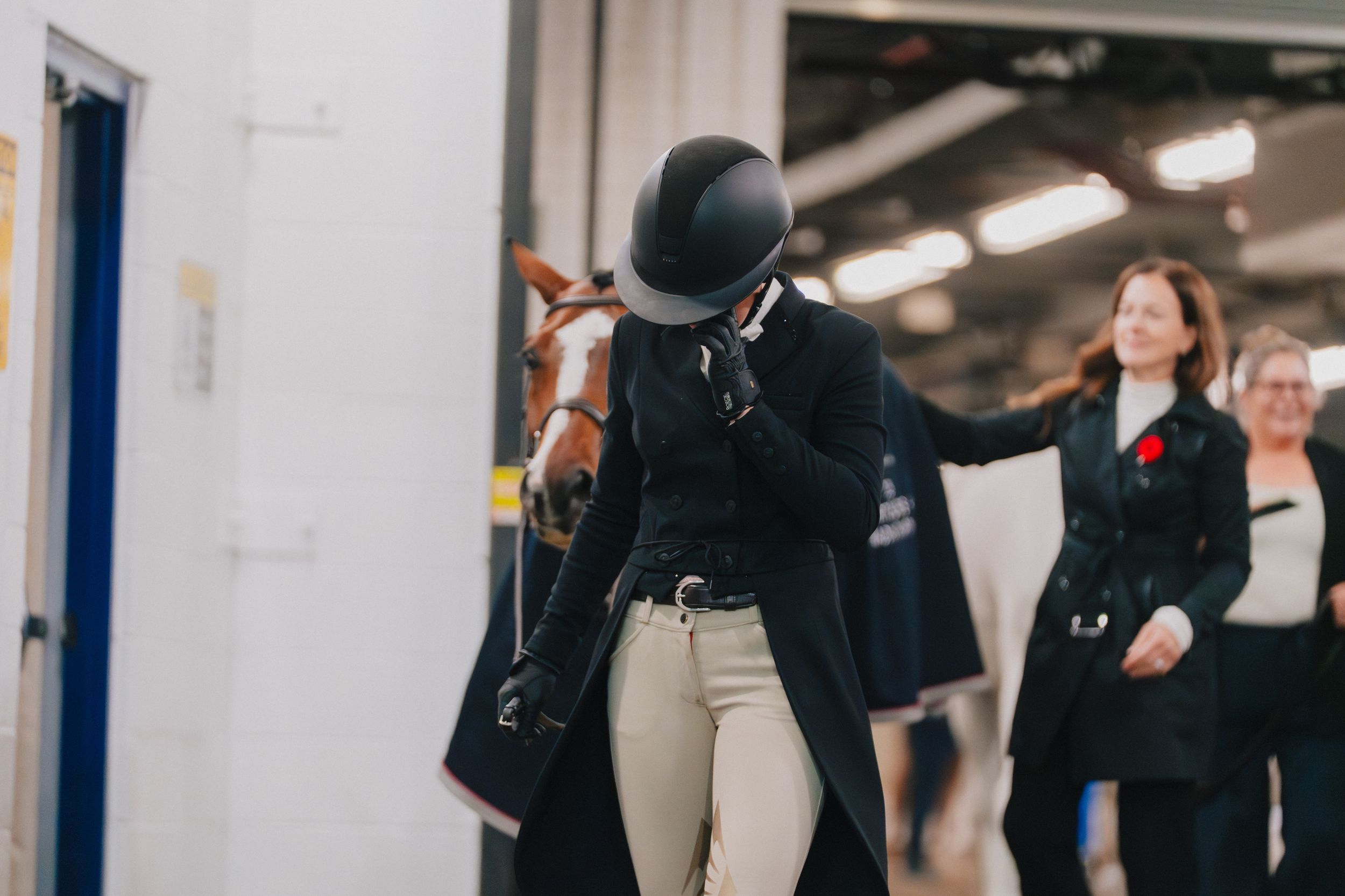 An equestrian in black show attire and helmet celebrates after a performance in an indoor arena.