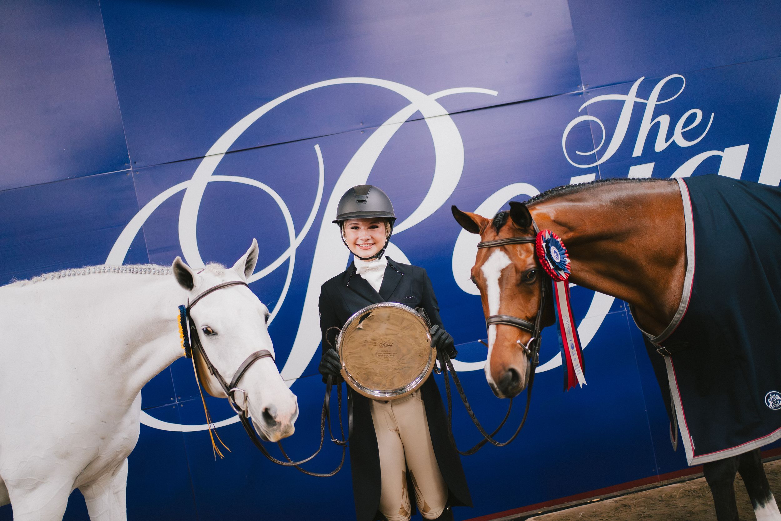 A rider poses with two horses, one white and one brown, in front of a blue branded backdrop.