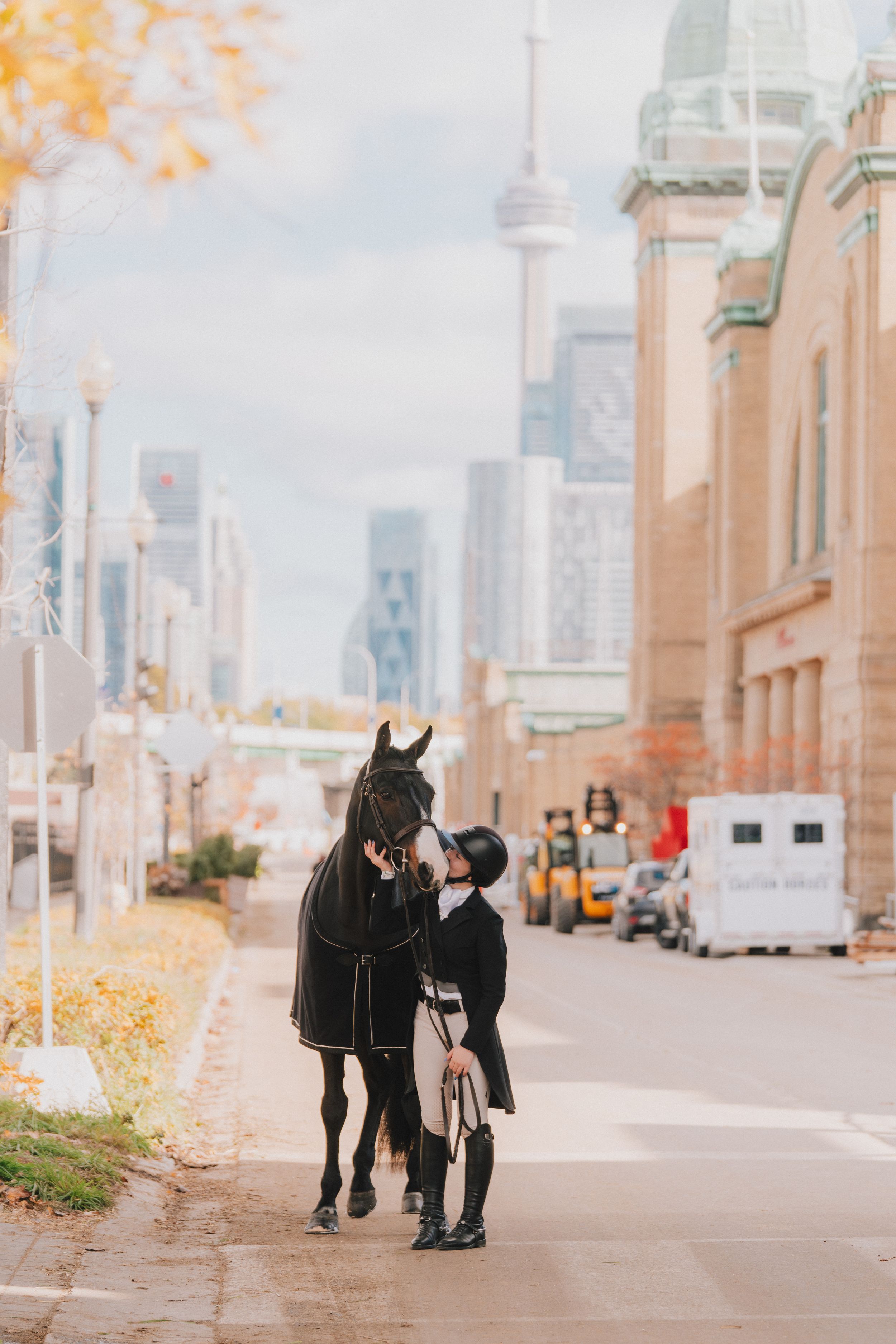 Two people walking together on a city sidewalk with tall buildings and autumn trees in the background.