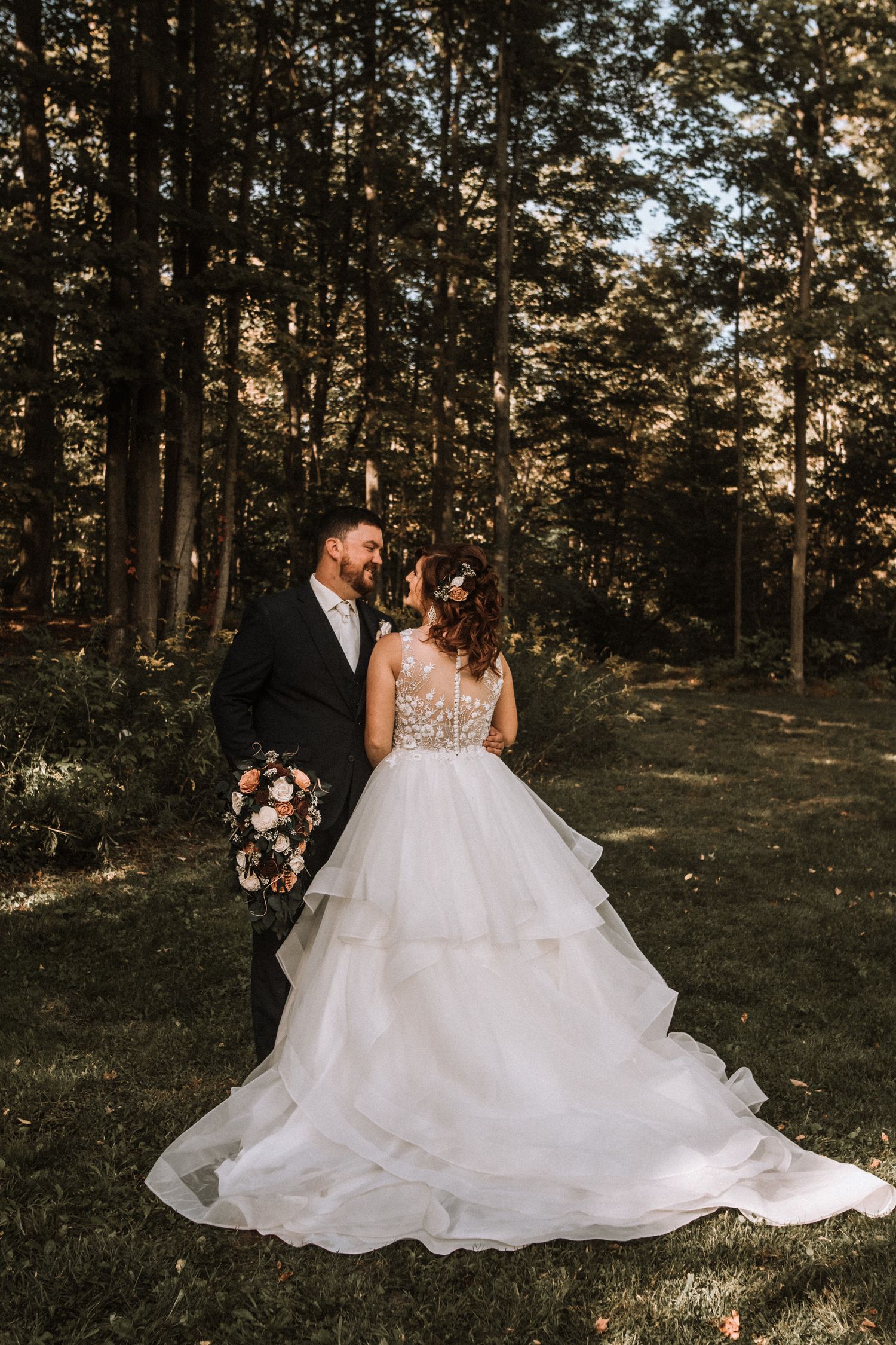 A couple shares a romantic moment in a sunlit forest wearing wedding attire.