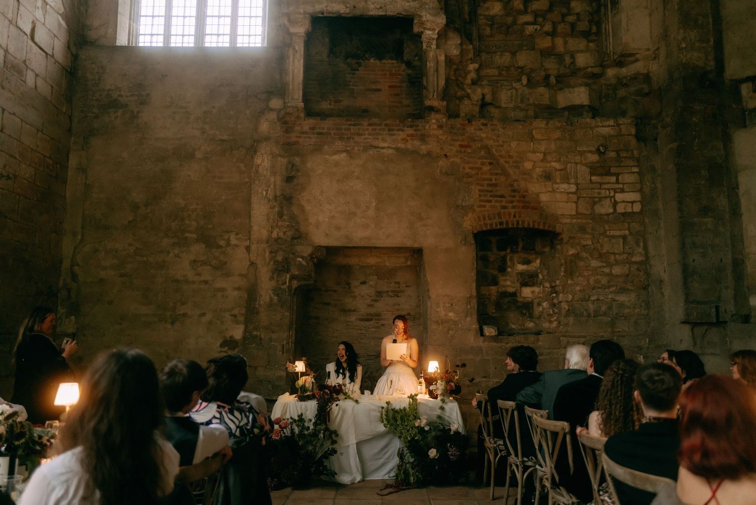 Candlelit wedding reception in a rustic stone castle with guests seated at long tables under glowing ambient lighting.