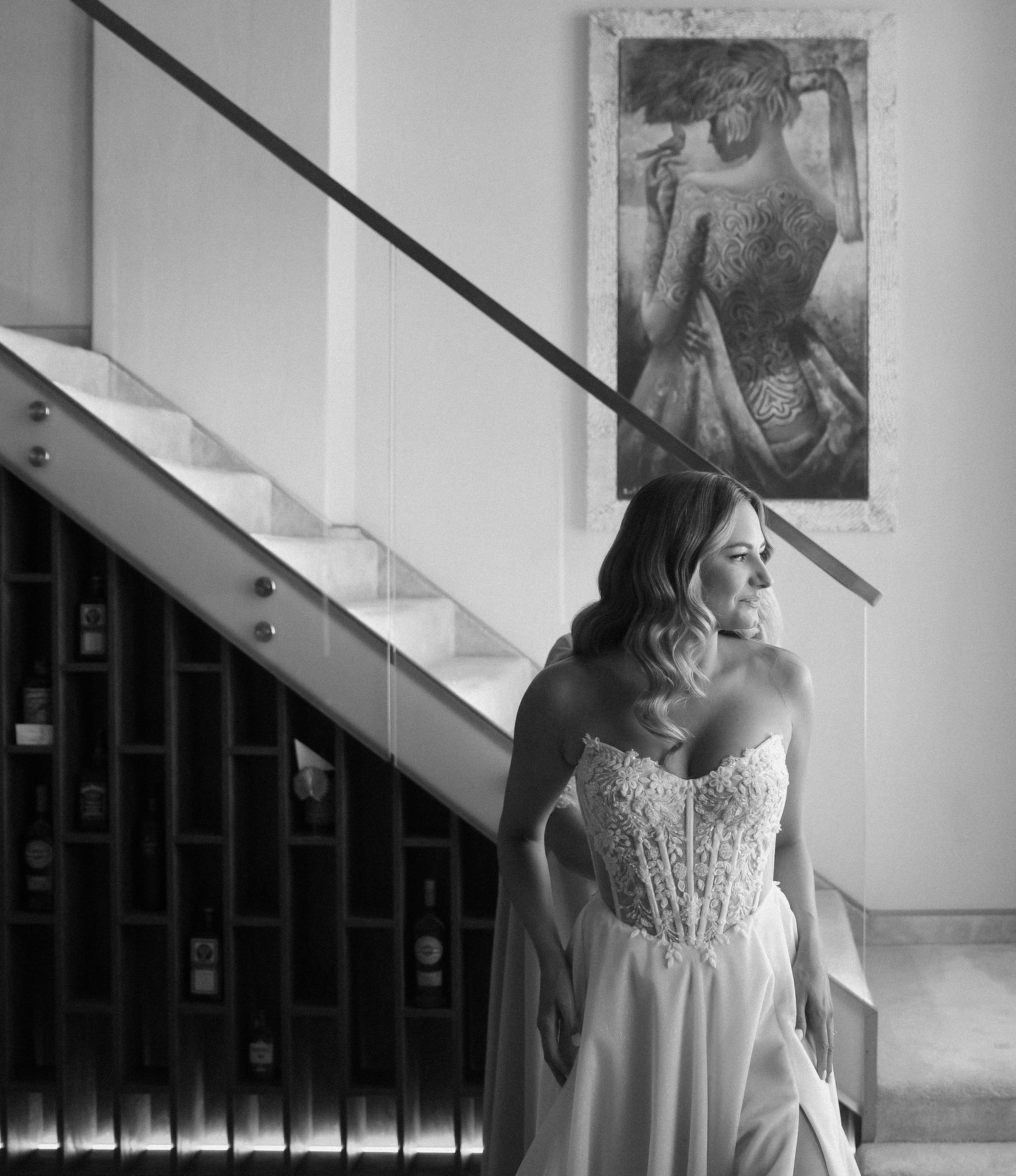 A bride in a white lace dress poses near a staircase in black and white photography.