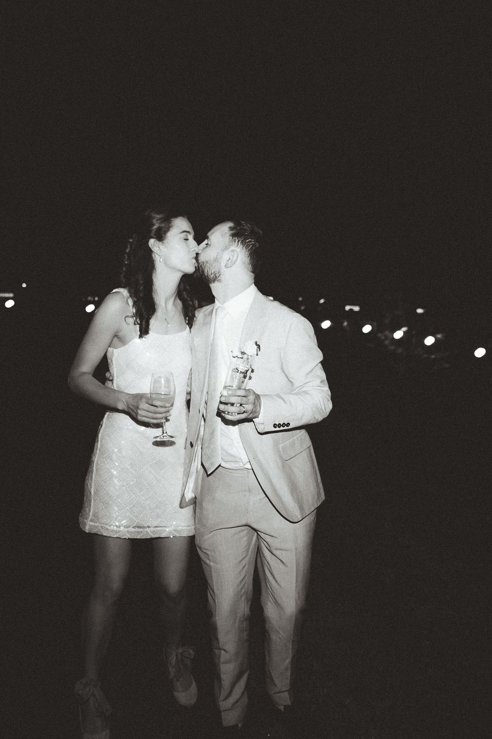 A black and white photo of a couple in formal attire sharing a romantic moment while holding drinks at night.