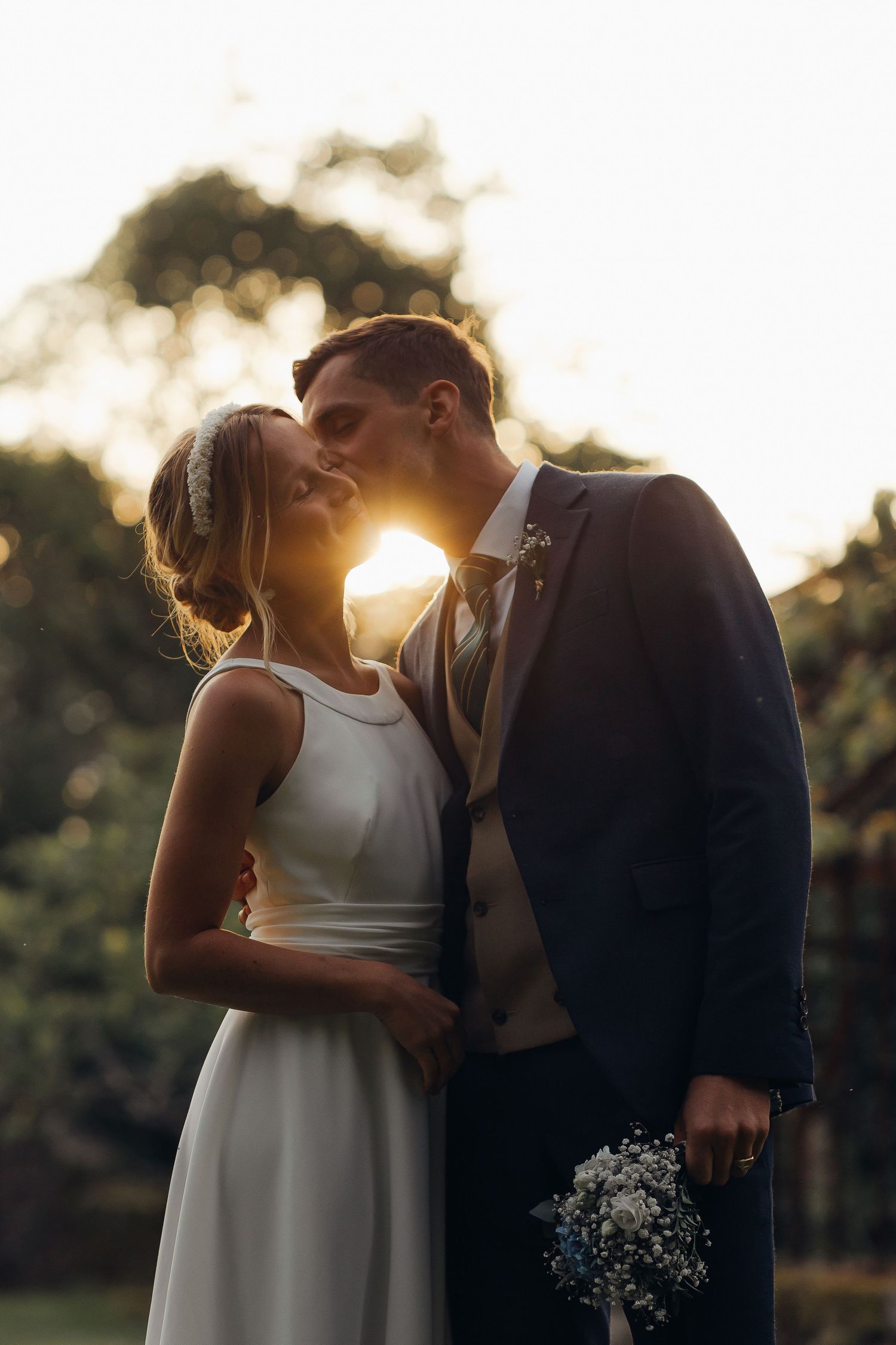 Bride and groom sharing a sunset kiss in garden, groom holding delicate wildflower bouquet.