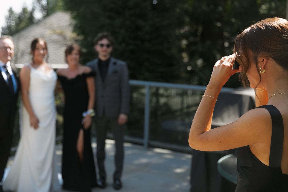 Wedding guest photographing bride, groom, and family members posing together outdoors on sunny wedding day.