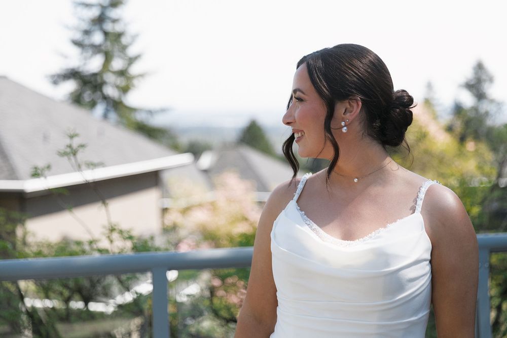 Beautiful bride in white gown with updo hairstyle and pearl earrings standing on outdoor balcony.