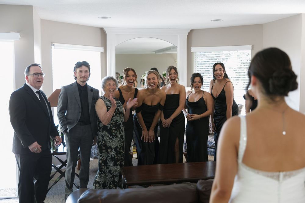 Bridal party in black dresses reacting to bride reveal indoors.