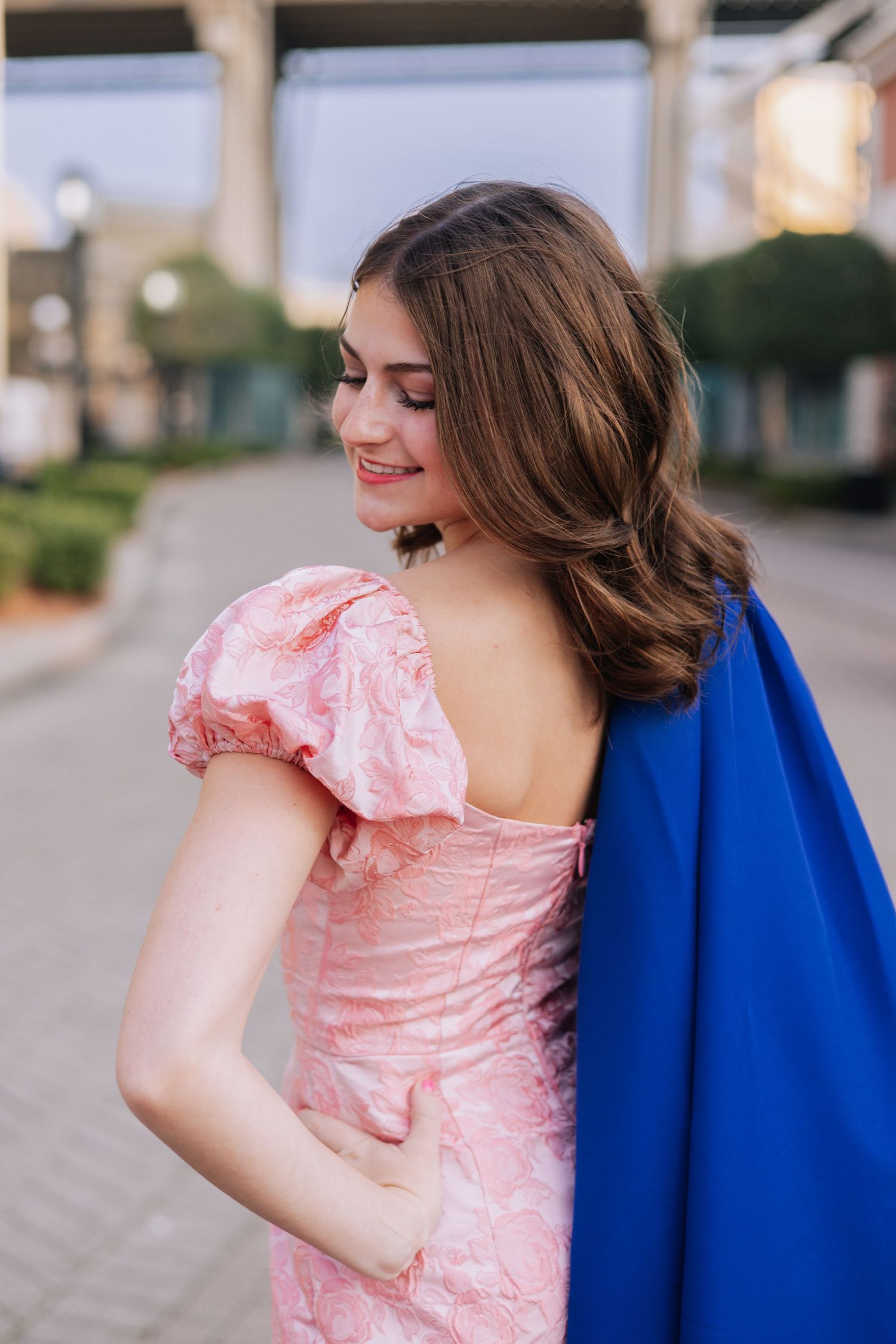 Woman in pink dress with blue cape twirls during outdoor maternity photoshoot.