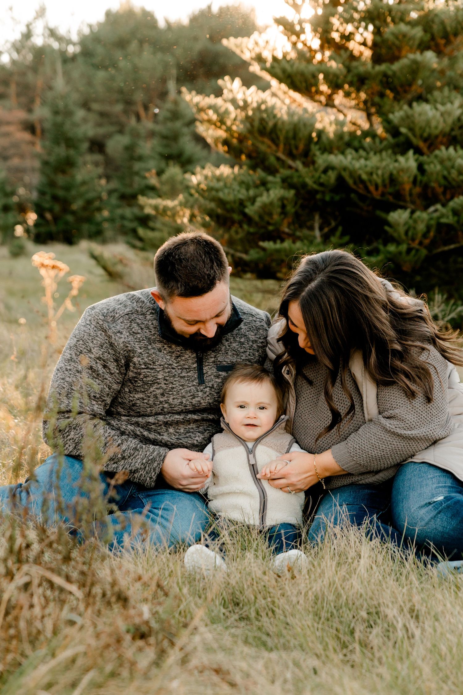 Family portrait of parents and baby sitting in golden grass field with evergreen trees in background.