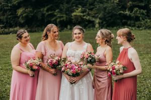 A group of bridesmaids in pink dresses stand together holding pink and red bouquets at an outdoor wedding.