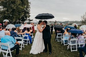 Outdoor wedding ceremony setup with white chairs and guests holding umbrellas on a cloudy day.