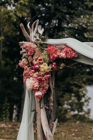 Rustic wedding arch decorated with pink and coral flowers, white fabric draping, and natural driftwood branches.